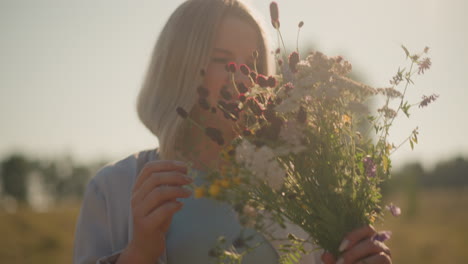 mujer sosteniendo un hermoso ramo de flores silvestres mientras las huele suavemente en el campo iluminado por el sol, el fondo presenta tonos dorados cálidos y vegetación distante