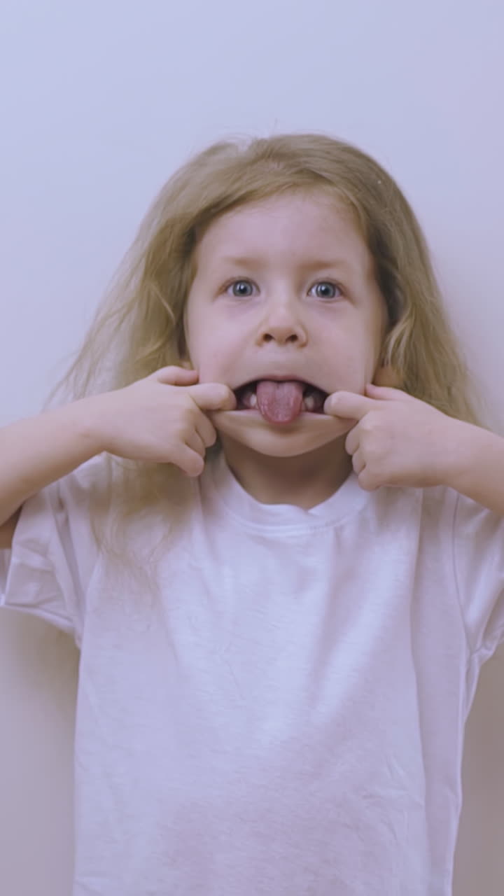 funny blonde little girl in white t-shirt makes face showing tongue at light wall in brightly lit room closeup