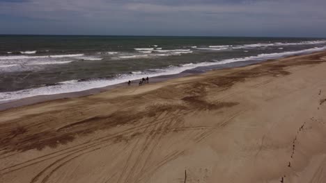 Toma-Aérea-De-Cuatro-Personas-Montando-Caballos-A-Lo-Largo-De-La-Playa-De-Arena-Con-Olas-Del-Océano-Atlántico-Durante-El-Día-De-Verano