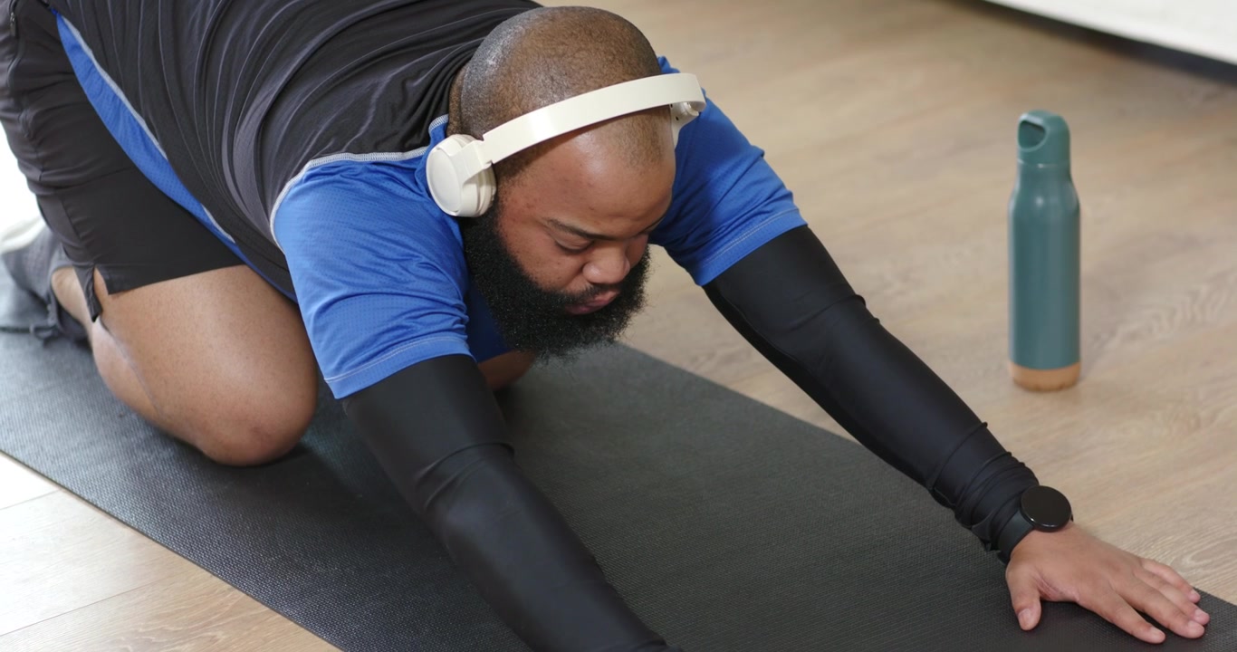 African American man wearing blue shirt stretching on yoga mat at home, lowering hips for fitness