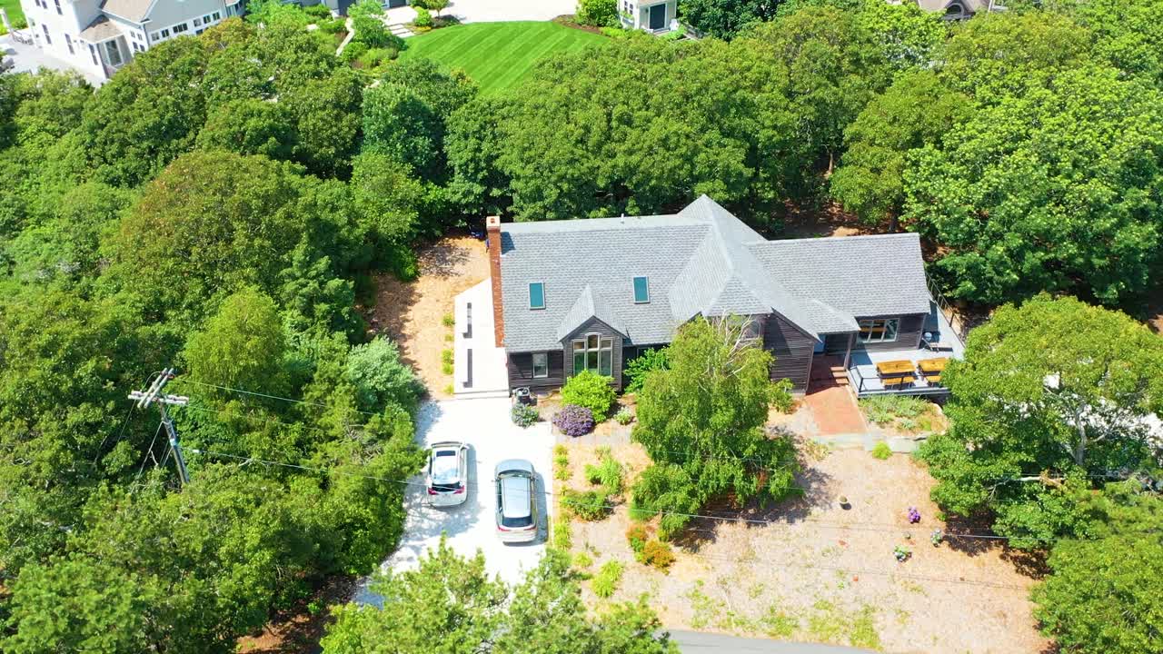 A gabled house with natural wood siding, wide windows, and shaded patio nestles into its wooded lot, revealed through an aerial rotation that highlights the home’s architectural detail forested charm