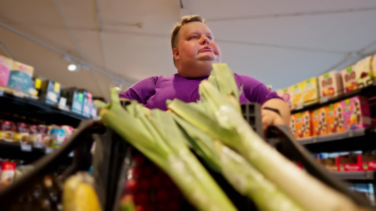 View from the cart side: A happy overweight man wearing a purple T-shirt walks through the supermarket, looks at the products and eats a croissant. Shallots are already in the man's cart