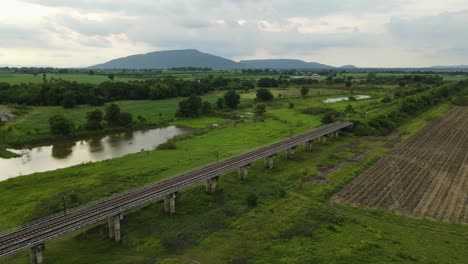 Imágenes-Aéreas-Hacia-Un-Hermoso-Paisaje-Y-Montañas-En-El-Horizonte-Que-Revelan-Un-Ferrocarril-Provincial-Elevado,-Estanques,-árboles-Y-Tierras-De-Cultivo,-Muak-Klek,-Saraburi,-Tailandia