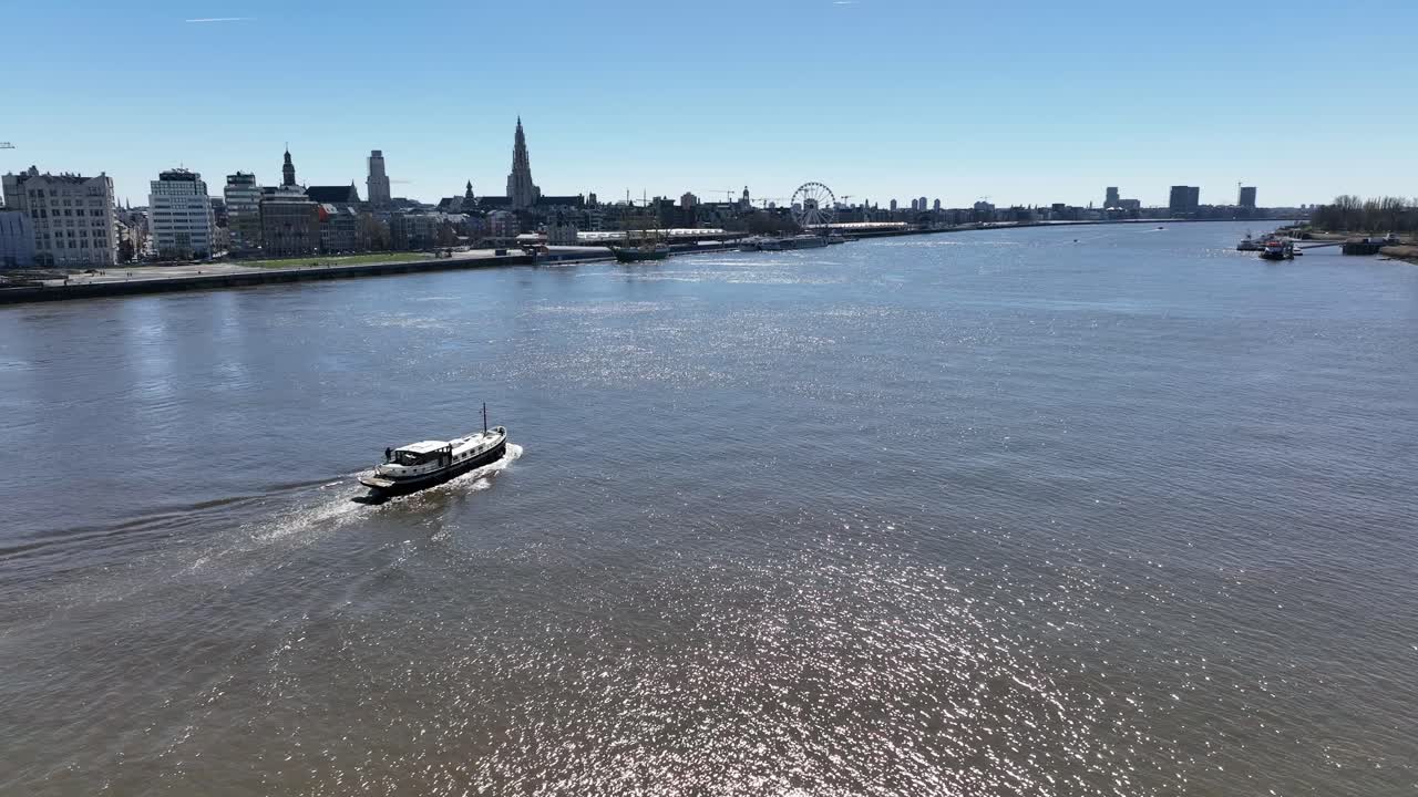 Semi orbit aerial shot of a boat cruising along the Scheldt river with Antwerp skyline and cathedral in the distance under bright daylight