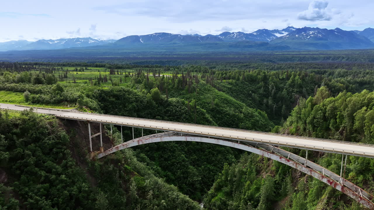 Connecting with Alaska beauty. A stunning view of a tall bridge spanning a lush canyon in Alaska, surrounded by majestic mountains and greenery