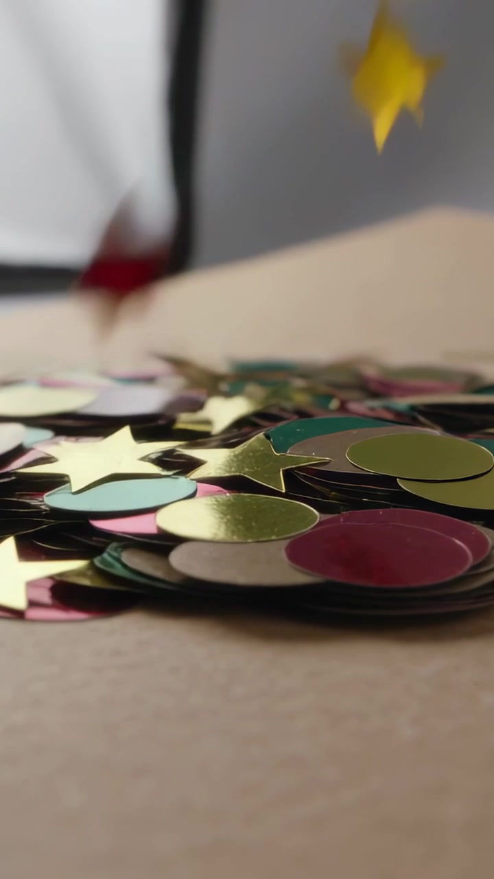 Vertical video: Falling metal confetti pieces hitting beige table, jostling pile with white panel