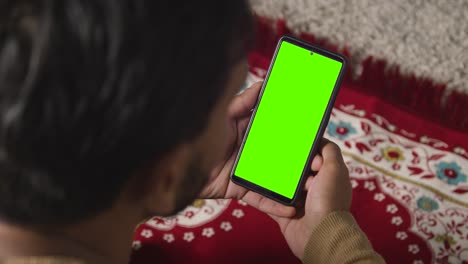 Close-Up-Of-Muslim-Man-At-Home-Sitting-On-Floor-On-Prayer-Mat-Holding-Green-Screen-Mobile-Phone-1