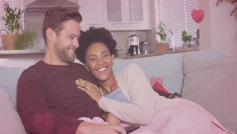animation of hearts over diverse couple embracing on sofa and watching tv