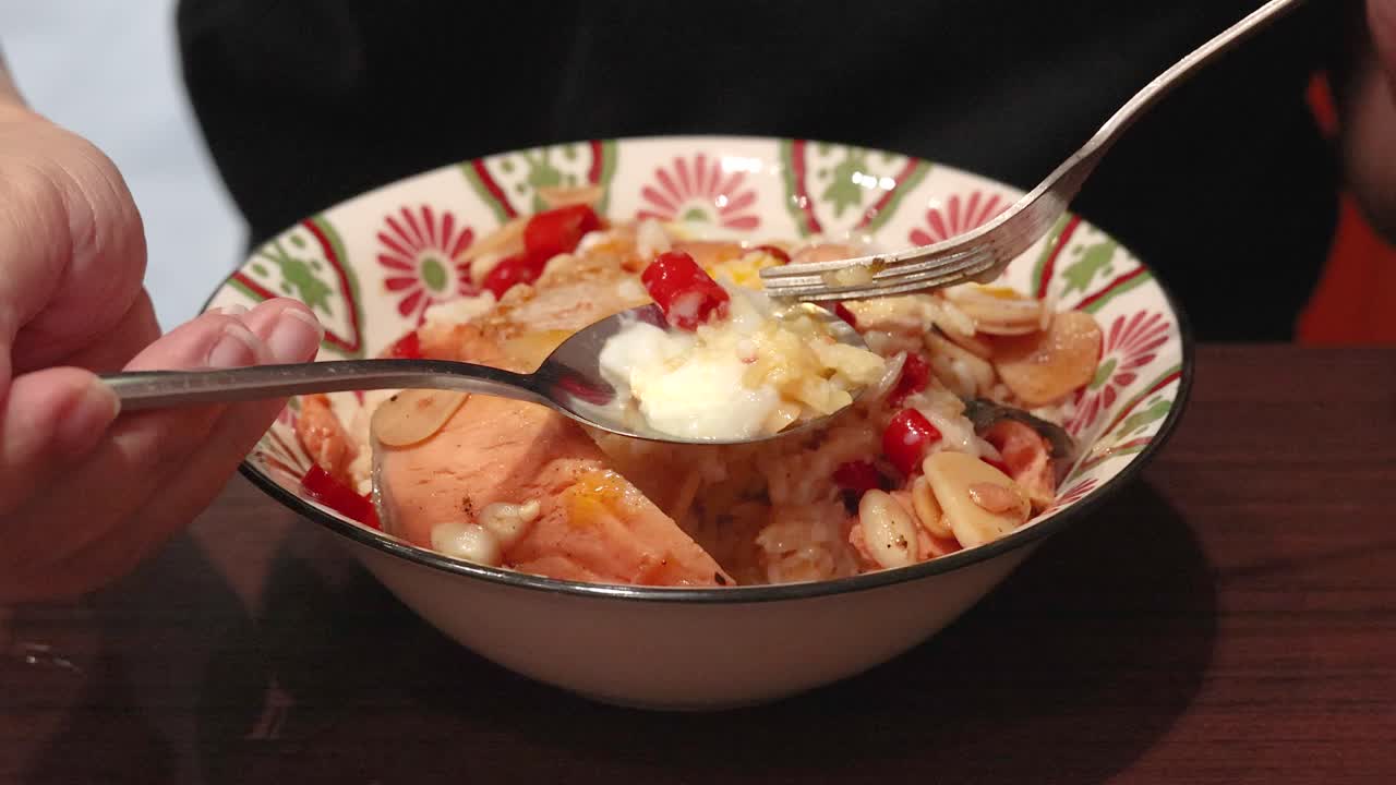 A person uses a spoon and fork to mix cooked salmon, rice, fried garlic, chili, and a soft egg in a patterned bowl under warm indoor lighting