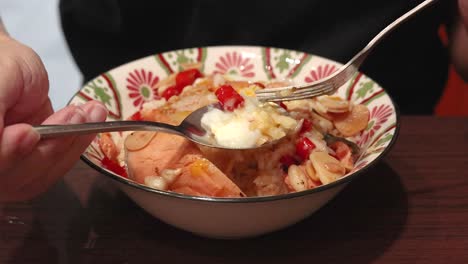 a person uses a spoon and fork to mix cooked salmon, rice, fried garlic, chili, and a soft egg in a patterned bowl under warm indoor lighting