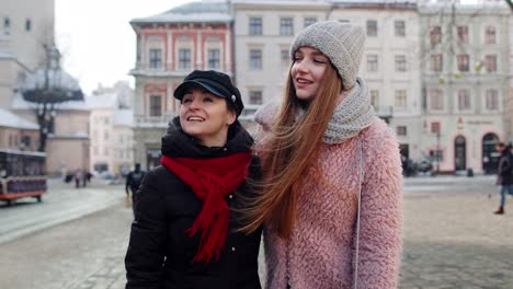 two women smile at each other while walking in a snowy european city.