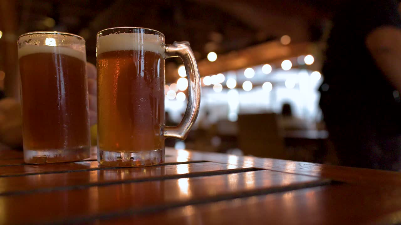 Due boccali di vetro freddi di birra lager ambrata sono su un tavolo di legno in un bar.