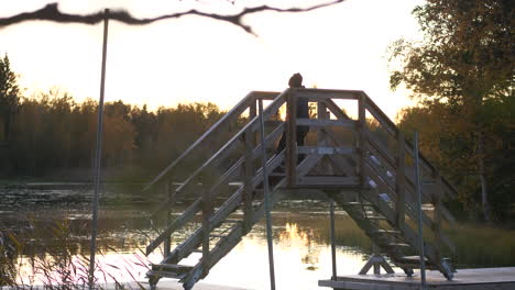 Lonely-young-boy-walking-over-a-small-wooden-bridge-at-dusk,-still-water-reflecting-the-last-sunrays