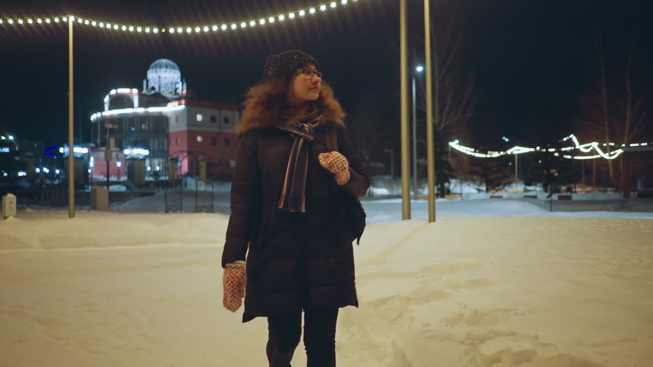 Woman in warm winter coat with scarf and colorful mittens walking on snowy path at night near illuminated building and festive lights, enjoying peaceful cold evening in cozy urban seasonal atmosphere