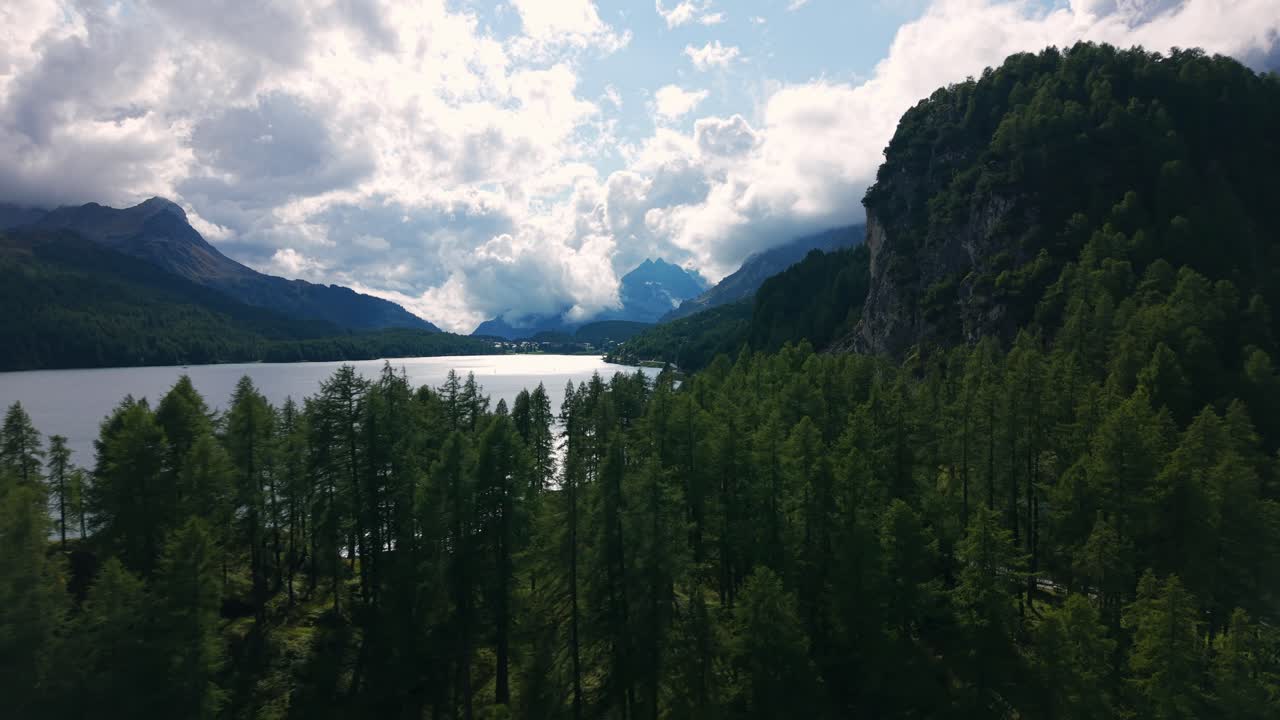 Silvaplana-søen, der reflekterer skyer, med grøn skov og bjerge under en lys himmel.