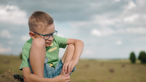 un niño joven se agacha en una bala de heno en una vasta tierra de cultivo, perdido en sus pensamientos, una escena rural iluminada por el sol captura un momento pacífico y reflexivo, con tonos cálidos de heno dorado en contraste con el campo abierto