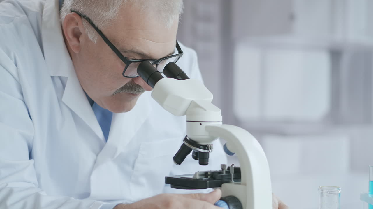 Bearded Caucasian male researcher wearing protective glasses and working with a microscope spbas. scientist using microscope in a laboratory. Search for coronavirus vaccine. High quality 4k footage