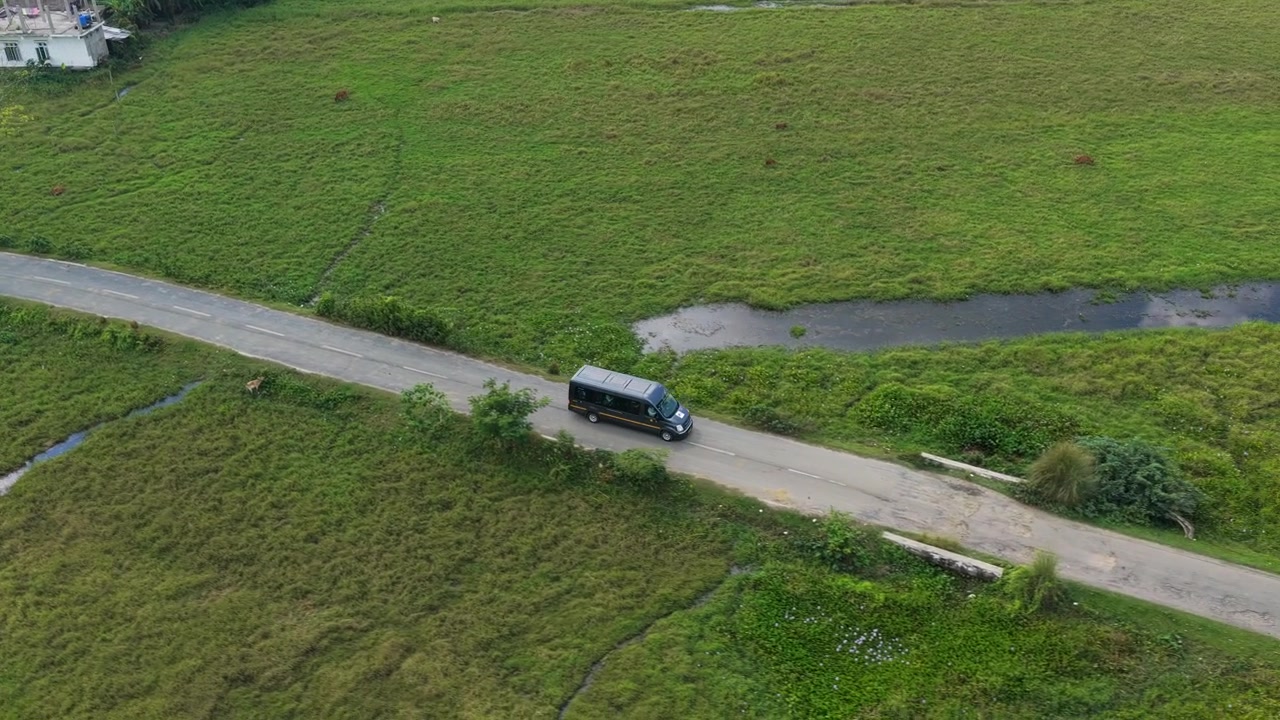Rekaman pelacakan udara sinematik sebuah van penumpang yang melaju melalui ladang pertanian hijau subur dan pemandangan pedesaan di India.