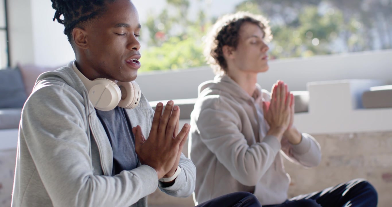 Two men sitting on balcony in morning sun, raising, folding hands, meditating, left wearing headset