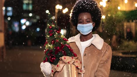 Close-up-view-of-joyful-African-American-woman-wearing-facial-mask-holding-a-Christmas-tree-and-smiling-at-camera-on-the-street-while-it¬¥s-snowing-in-Christmas