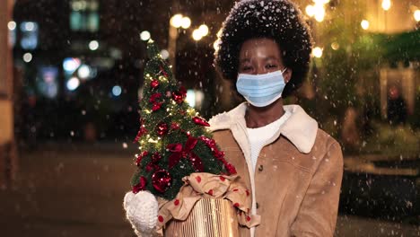 vista de cerca de una alegre mujer afroamericana con máscara facial sosteniendo un árbol de navidad y sonriendo a la cámara en la calle mientras nieva en navidad