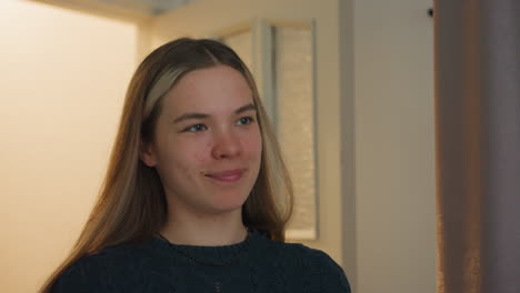 blonde woman wearing black knitted sweater and subtle necklace walks through white wooden door into softly illuminated hallway, with geometric wooden wall accents and frosted glass panel
