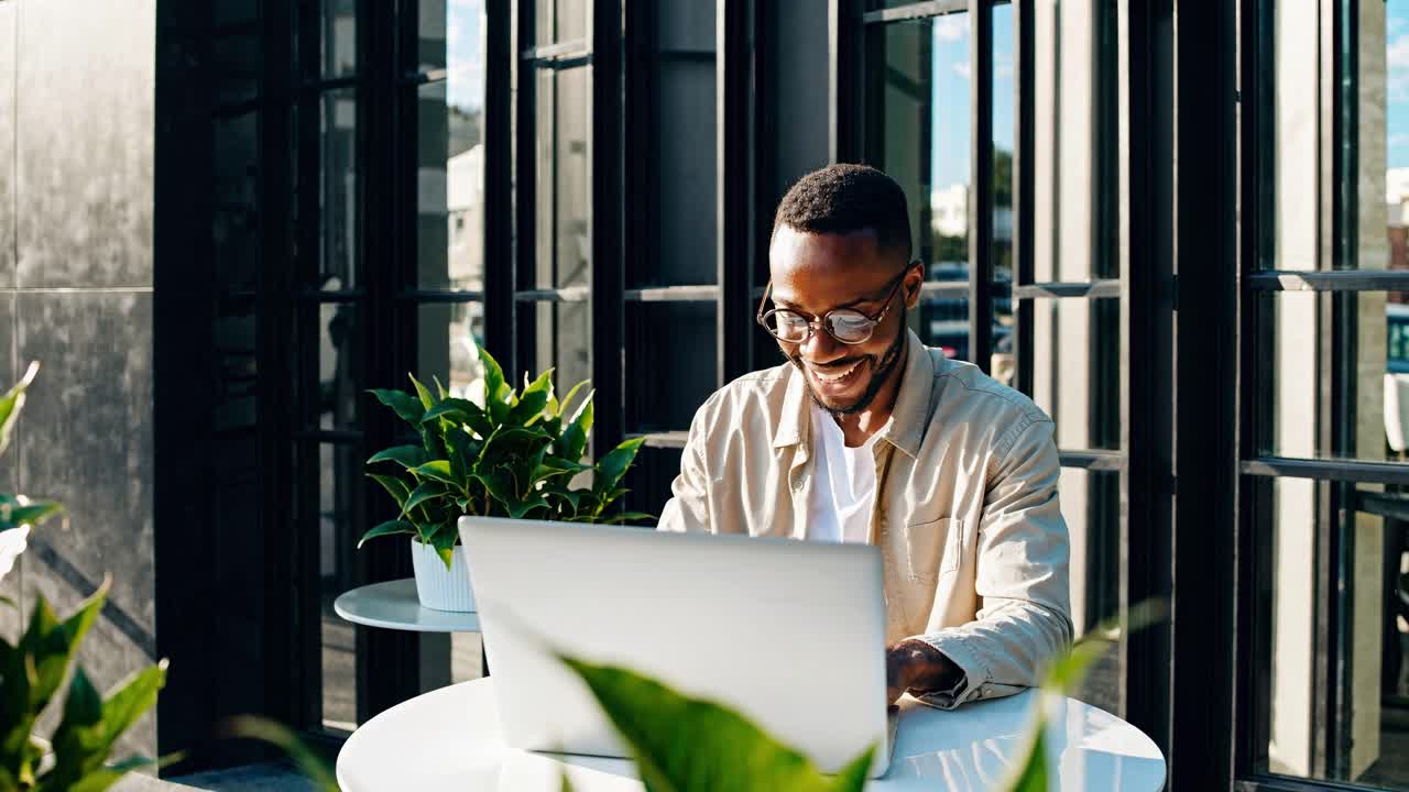 Un uomo che sorride mentre lavora al laptop in un caffè all'aperto. L'inquadratura dal basso cattura una scena luminosa.