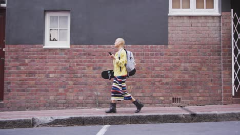 Mixed-race-woman-walking-on-street-holding-her-skateboard-and-her-phone-