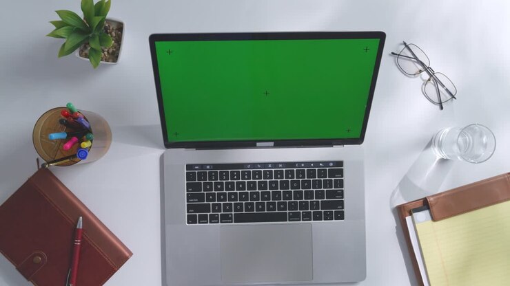 Draufsicht auf einen Laptop-Computer mit Mockup-Greenscreen-Display auf einem Holzschreibtisch, daneben ein Notizbuch mit Stiften, Brille und einem Glas Wasser. Langsames Herauszoomen, Nahaufnahme.