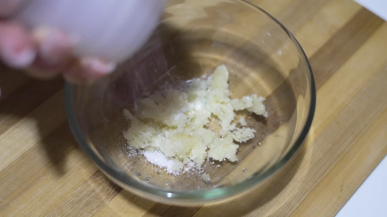 Close up of crushed garlic mixed with salt in a glass bowl, ready for cooking preparation.