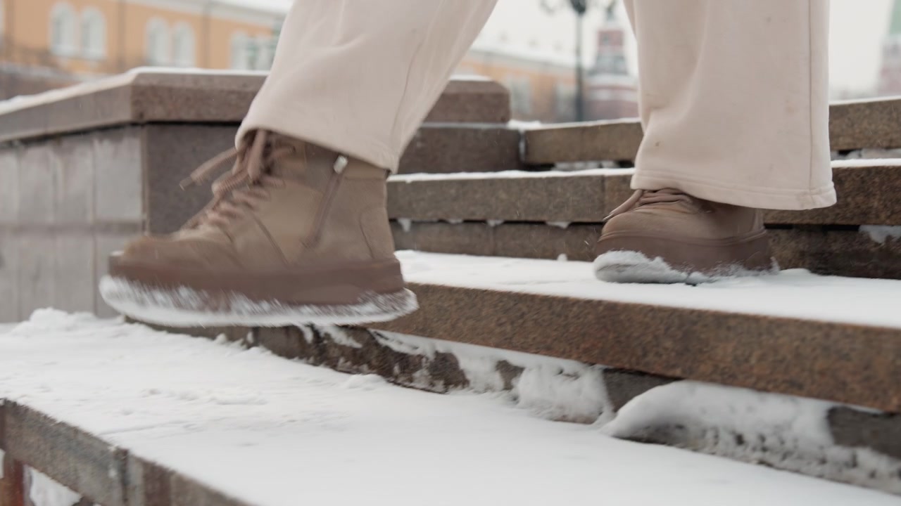 Low angle view shows person descending snow covered stone steps in urban winter setting, brown shoe meeting textured tread, light pant leg in motion, careful stride, cold overcast daylight, city
