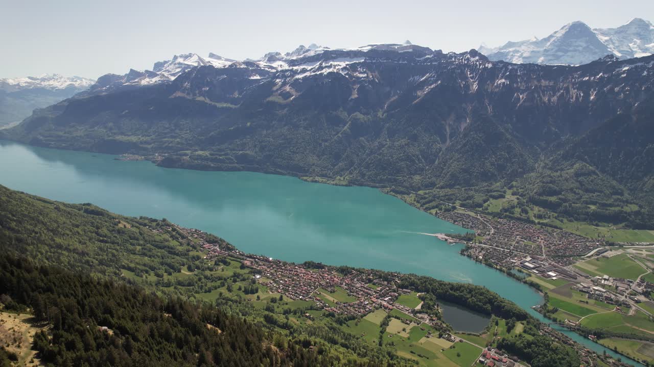 Drone shot above Lake Brienz, Interlaken and the Swiss Alps. Clear turquoise water, alpine villages and snow-covered peaks under spring sunlight
