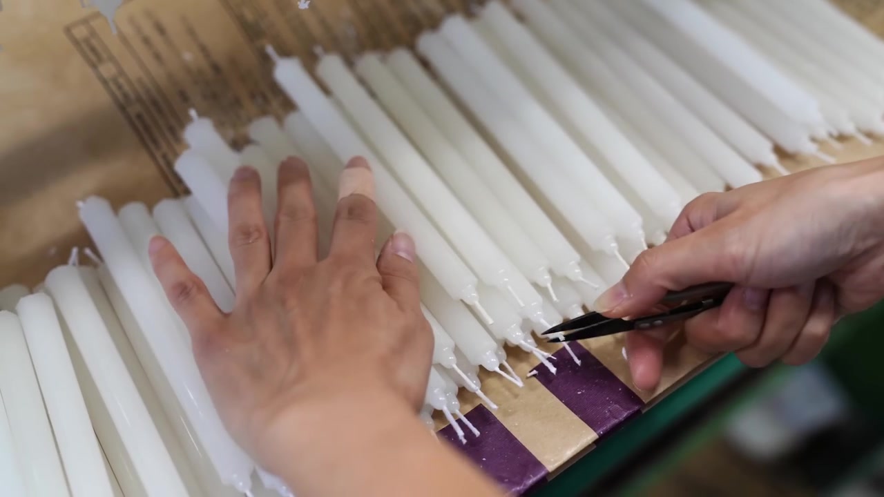 Werknemer die lonten trimt aan de basis van taps toelopende kaarsen, liggend op een met papier bedekte fabriekstafel.