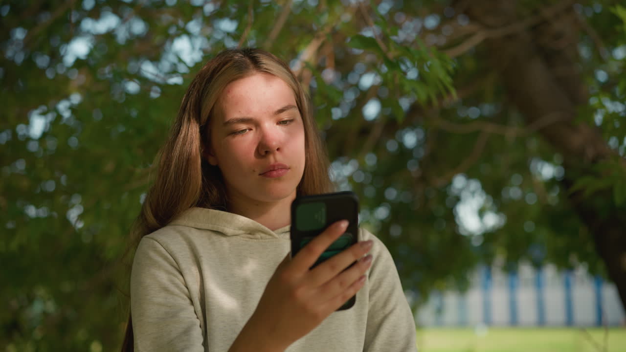 Young woman focused on her phone under a tree with sunlight filtering through leaves, creating soft shadows on her face, she appears engaged in her screen