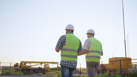 Oberkörperaufnahme-Von-Zwei-Männlichen-Bauarbeitern-Mittleren-Alters-In-Sicherheitskleidung,-Die-Auf-Der-Baustelle-Stehen.-Mann-Benutzt-Walkie-Talkie,-Sein-Kollege-Hält-Papier-Mit-Projektplan-In-Der-Hand