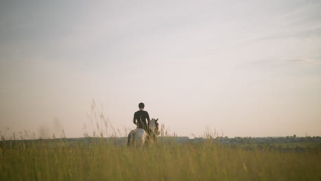 a peaceful, back-facing shot of a person dressed in black, riding a horse through a vast grass field during sunset. the warm, fading light of the day casts a serene glow over the landscape