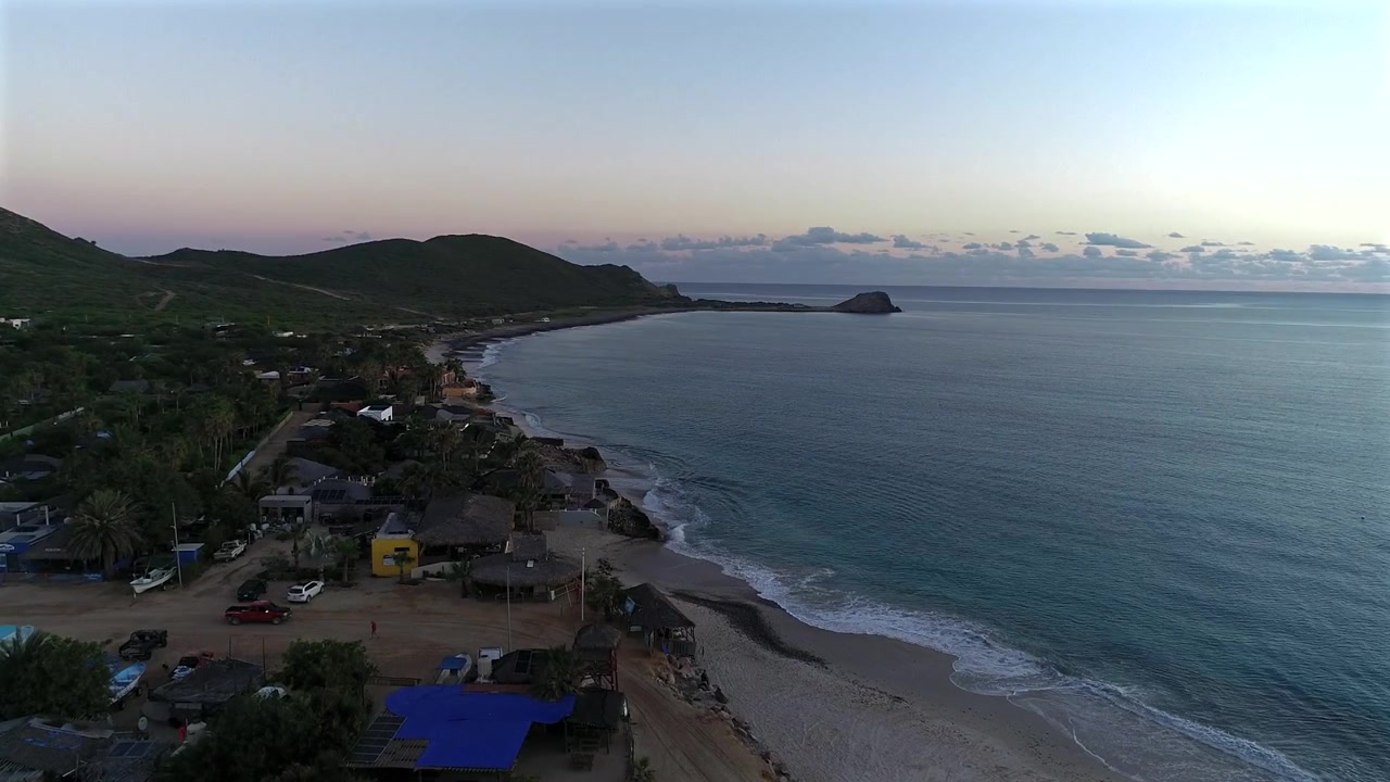 Un drone survole la pointe de Cabo Pulmo en Basse-Californie du Sud, se déplaçant vers l'intérieur des terres avant de tourner la caméra vers l'océan, surplombant la plage principale tandis qu'un camion s'arrête le long du rivage