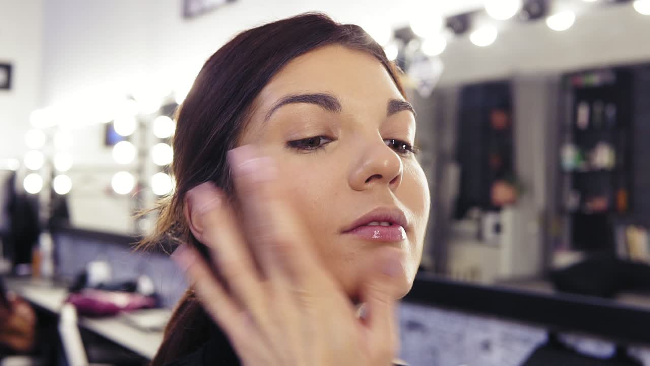 Process of make up artist's hand applying foundation on previously prepared face of an attractive brunette girl in a beauty salon or studio.