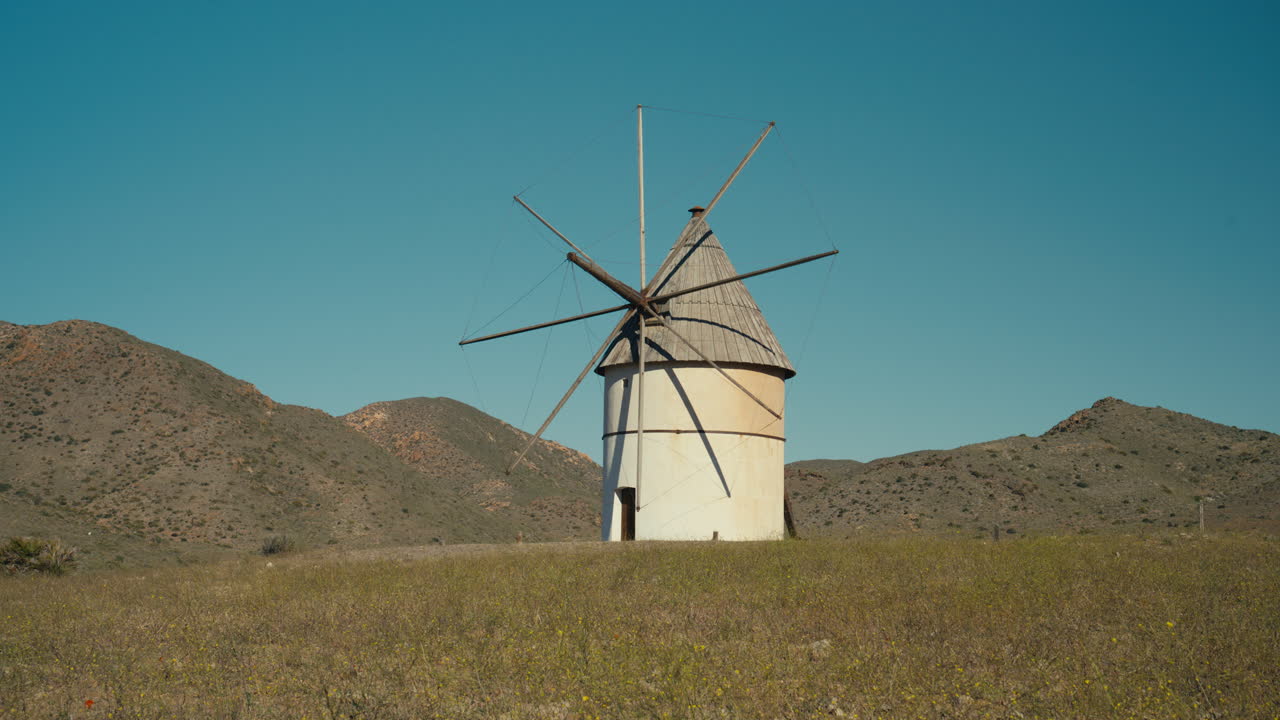 Molino de viento tradicional solitario en un paisaje mediterráneo seco bajo un cielo azul claro. Destino turístico pintoresco con arquitectura histórica y un entorno rural tranquilo.