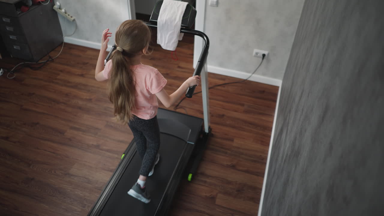 Rear view of young girl stepping onto treadmill holding handles hair tied back in ponytail to begin workout session in home fitness room with shelf electric socket and grey textured wall wooden floor