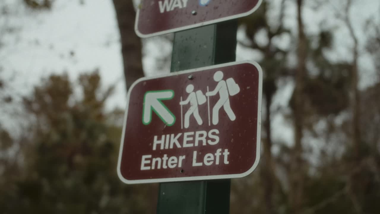 A close-up shot of a brown and white hiker entrance sign in a wooded area. The sign, marked with an arrow and two backpacking figures, directs hikers to enter left.