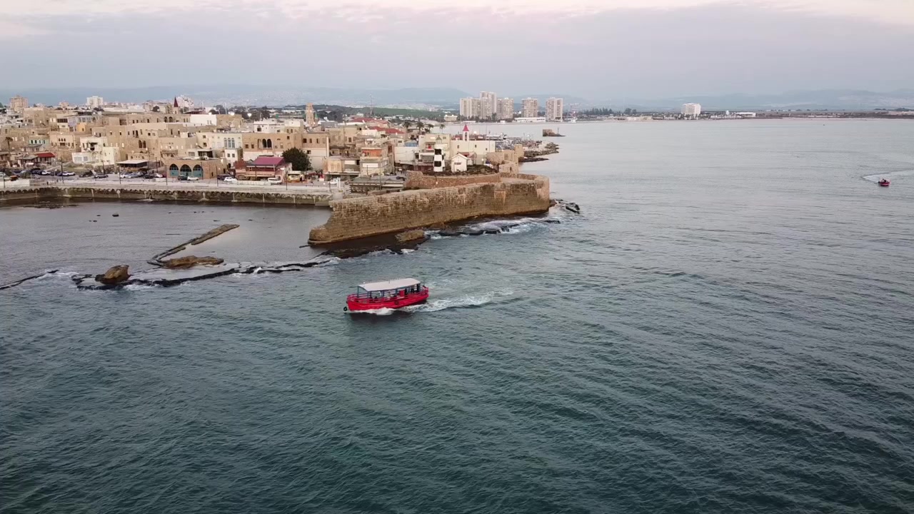 Riprese aeree al crepuscolo di una piccola barca rossa che attraversa le antiche mura marittime e il patrimonio architettonico di Akko, Città Vecchia di Acri in Israele.