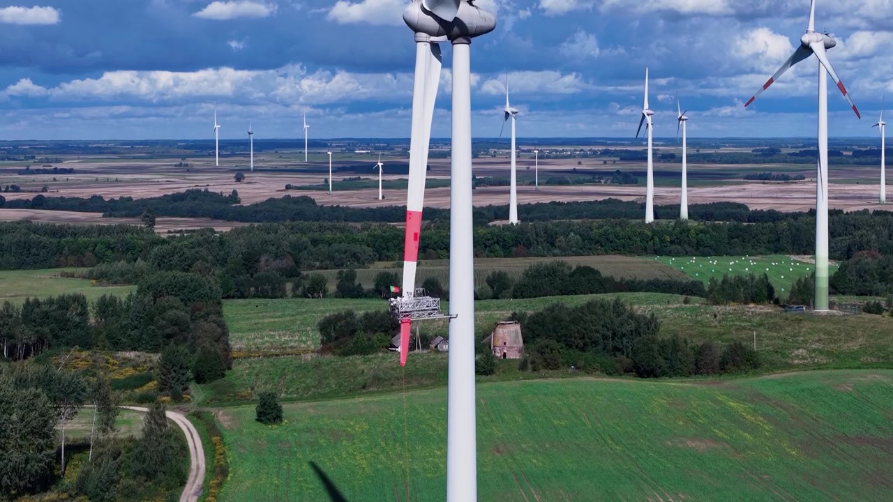 Flyfoto viser en tekniker som utfører vedlikehold på toppen av en vindturbin omgitt av store grønne felt, under dramatiske himmelstrøk, som fanger bærekraftig energi i aksjon