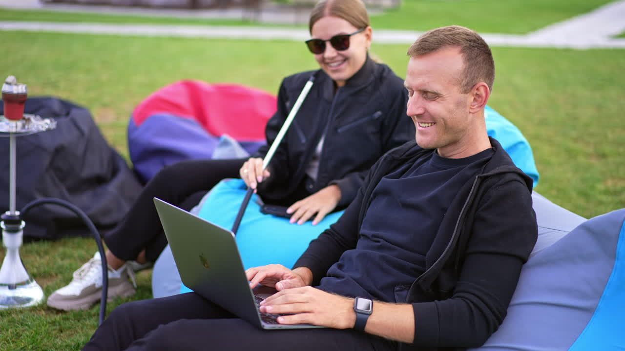 Smiling happy young people sitting in bean bag chairs on green grass. Man using laptop and woman smoking hookah.