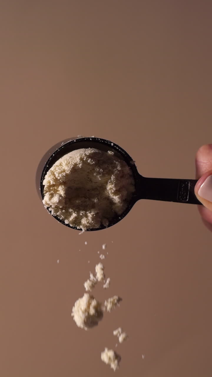 Slow-motion close-up of chocolate or vanilla protein powder gently falling from a black scoop against a warm beige background. Fine particles float midair, emphasizing texture, purity, and motion.