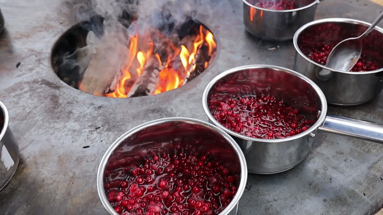 Several stainless steel pots filled with red berries simmer on a flat metal surface above an open fire pit, with steam rising from the bubbling mixtures