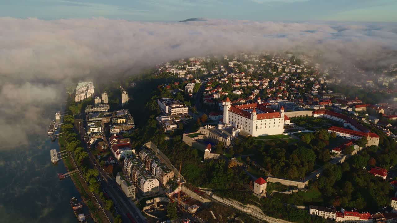Aerial shot of Bratislava Castle and Slovak Parliament in Slovakia during sunrise, partially covered by morning fog. Historic architecture and residential area with vibrant rooftops