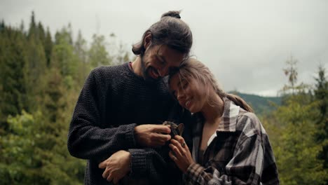 A-guy-in-a-blue-sweater-and-a-blonde-girl-are-stroking-a-cat,-which-is-being-held-by-a-brunette-guy.-Rest-with-a-pet-in-a-country-house-overlooking-a-coniferous-mountain-forest