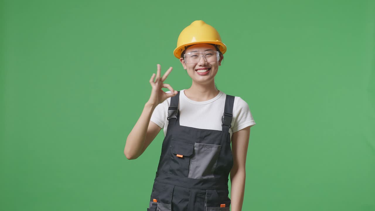 안경과 안전 헬을 착용 한 아시아 여성 노동자가 미소 짓고 녹색 화면 배경 스튜디오에 서있는 동안 카메라에 은 몸짓을 보여주고 있습니다.