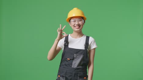 asian woman worker wearing goggles and safety helmet smiling and showing okay gesture to camera while standing in the green screen background studio